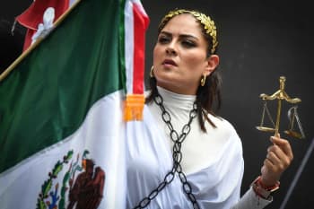 A woman wearing a white dress, holding a Mexican flag in one hand and a scale of justice in the other, with a chain draped around her neck. In the background, two other people can be seen, and the background is dark.
