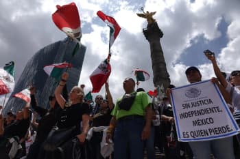 A group of people holding Mexican flags in front of a statue, with buildings in the background and clouds in the sky. Some of the people are wearing caps and holding placards with text, while others are holding mobile phones. The scene is likely related to Mexico's independence day, as indicated by the text on the placards.
